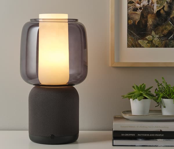 SYMFONISK bordlampe med wi-fi-højttaler i hvid/grå på et sengebord ved siden af en seng.