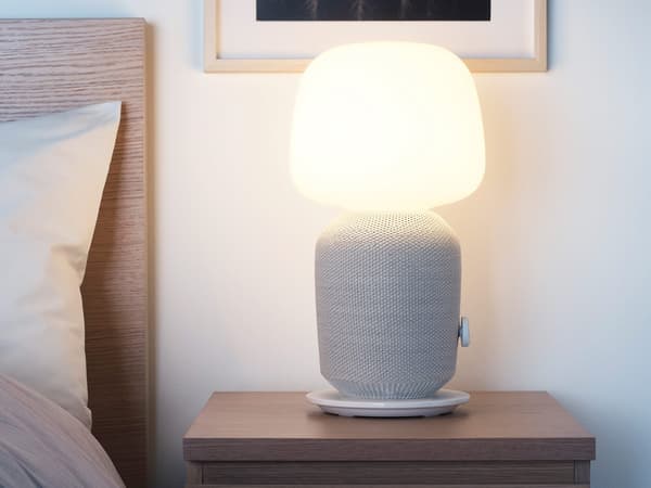 SYMFONISK table lamp with WiFi speaker in white/grey placed on a bedside table next to a bed.