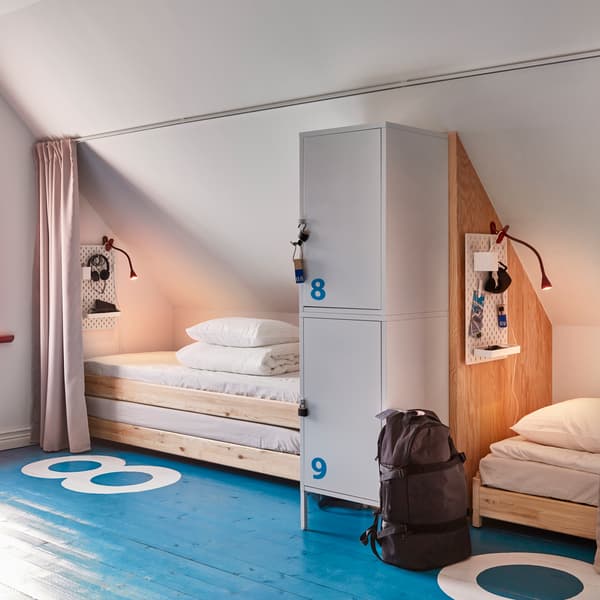 Sypialnia ze skośnym sufitem i niebieską podłogą z numeracją wskazującą dwa oddzielne miejsca do spania, wyposażone w drewniane łóżka UTÅKER i zamykane na kłódkę szafki.