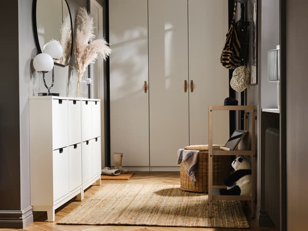 Szafki na buty STÄLL stoją pod dużym okrągłym lustrem, a obok leży dywan LOHALS z juty. Rząd białych szaf PAX ustawionych pod przyległą ścianą.