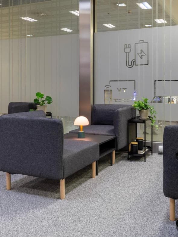 Szara kanapa stoi na szarej wykładzinie przy przeszklonej ścianie.