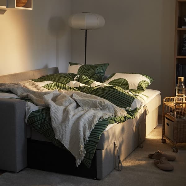 Szara sofa rozkładana z pościelą stojąca w słabo oświetlonym pokoju dziennym.