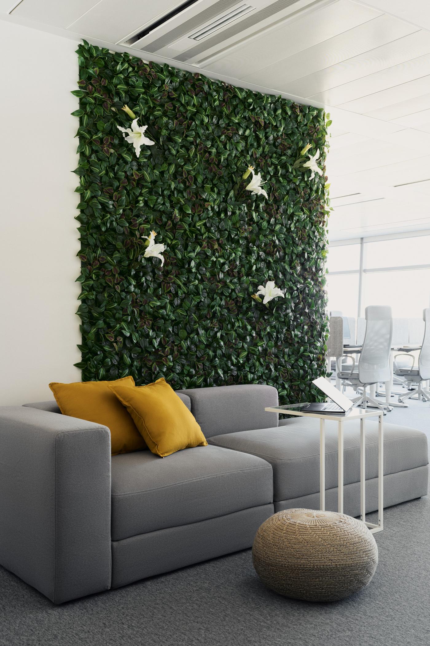 Szara sofa z żółtymi poduszkami, puf i zielona ściana z białymi kwiatami w biurze.