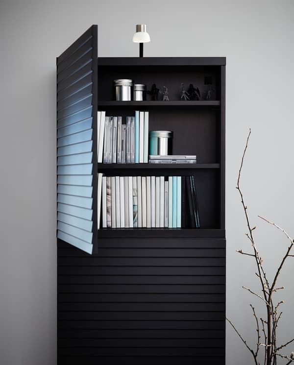 Szekrény hangsúlyos, kidomborodó antracit rácsos ajtókkal és tetején nikkelezett LED lámpa. Egy ajtó nyitva van, és könyvek vannak benne.