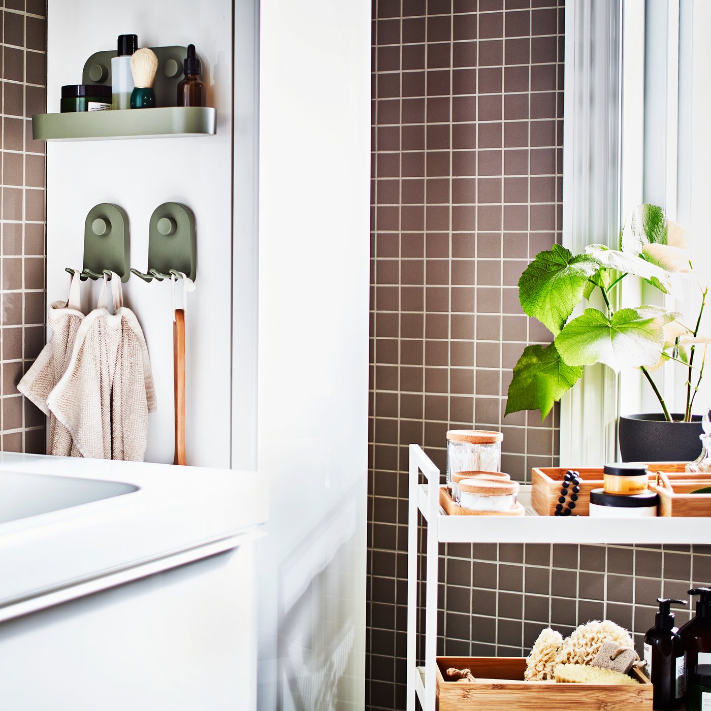 Szürke csempével burkolt fürdőszoba fehér mosdópulttal, fehér magasszekrénnyel, tálcákkal és üvegekkel megrakott, fehér zsúrkocsival, valamint egy cserepes növénnyel.