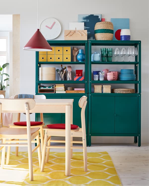Table and chairs in birch, red chair pads, a yellow rug, a dark red pendant lamp and two green shelving units with cabinets.