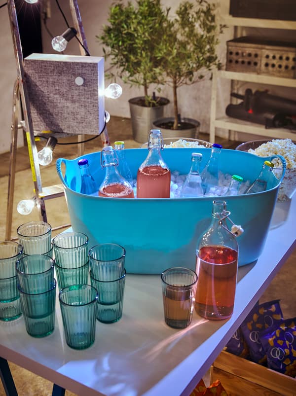 Table avec verres KALLNA empilés à côté d’un panier à linge TISKEN rempli de glace et de boissons dans des gobelets fermés, y compris des bouteilles en verre KORKEN.
