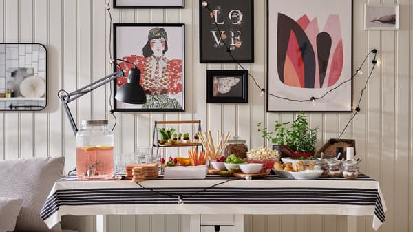 Table de buffet avec des plats et boissons dans différents bols, de la vaisselle et une fontaine à eau VARDAGEN, et un mur avec des images encadrées.