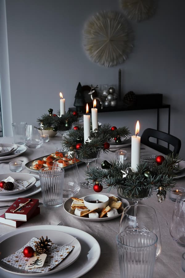 Table de fête avec des bougies blanches allumées, des centres de table à feuilles persistantes, des décorations rouges et une vaisselle élégante. Ambiance chaleureuse et accueillante.