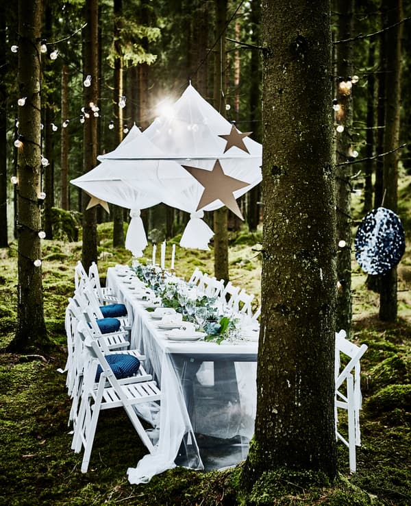 Table de noces dans la forêt avec longue table blanche, chaises pliantes blanches, objets décoratifs suspendus et guirlandes lumineuses enroulées autour d’arbres