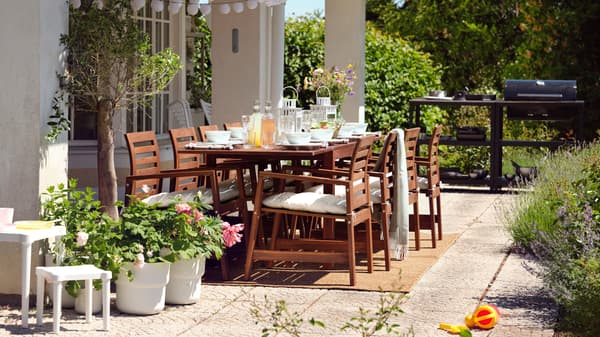 Table en bois ÄPPLARÖ et huit chaises sur une terrasse garnie de plantes, barbecue GRILLSKÄR à côté et guirlande lumineuse au-dessus.