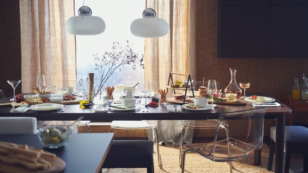 Table extensible STRANDTORP devant une fenêtre, dressée pour un grand repas avec assiettes, verres, fleurs et nourriture.
