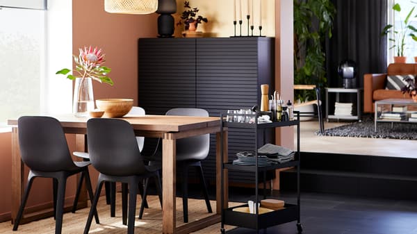 Table MÖRBYLÅNGA en chêne plaqué avec des chaises ODGER noires près d’une fenêtre, avec une armoire noire et une desserte contenant de la vaisselle.