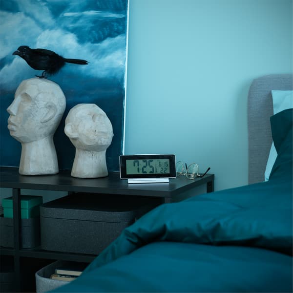 Table noire portant un réveil et des articles déco près d'un lit aux textiles vert foncé.