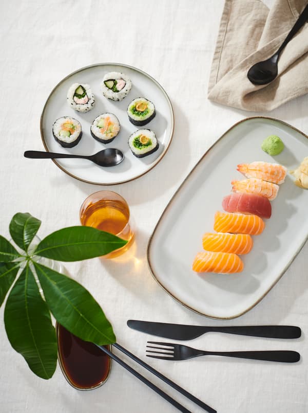 Table section with white tablecloth, sushi served on GLADELIG plates, TILLAGD cutlery and a decorative plant twig in a glass.