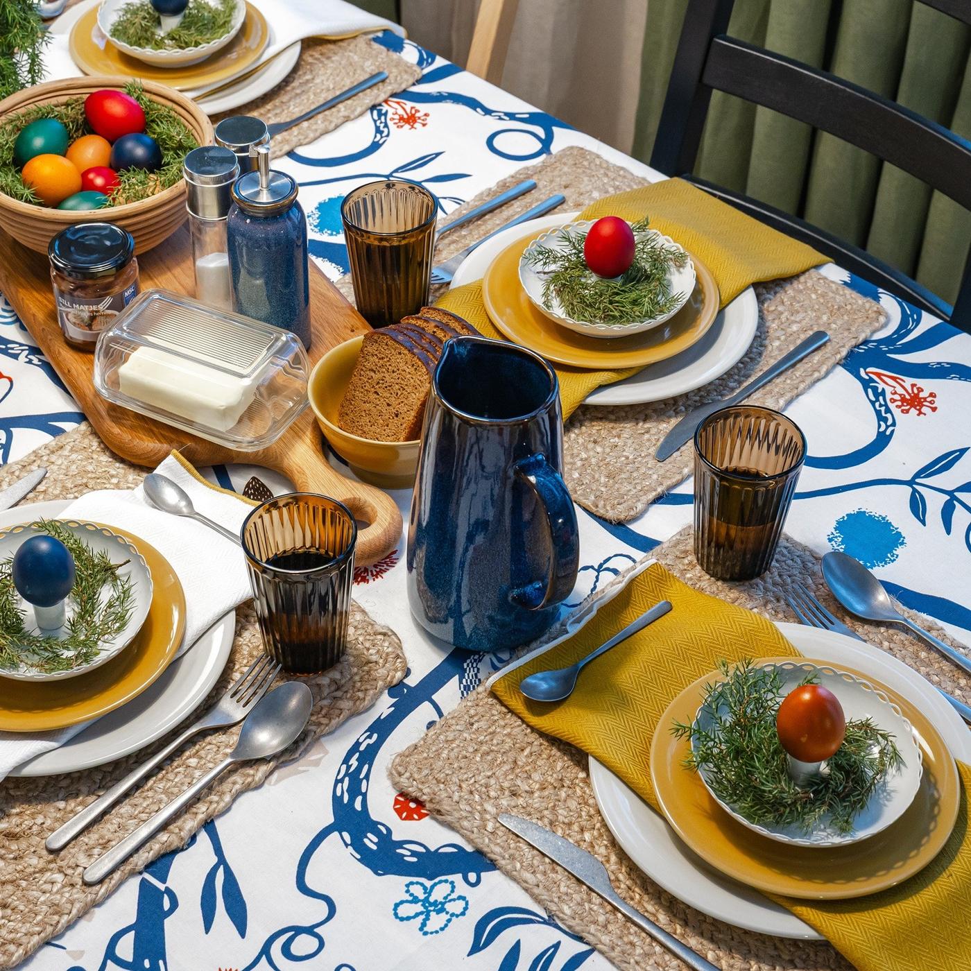 Table set with a vibrant precut fabric featuring blue floral motifs, styled with amber glasses, blue jugs and colourful eggs as playful decorations.