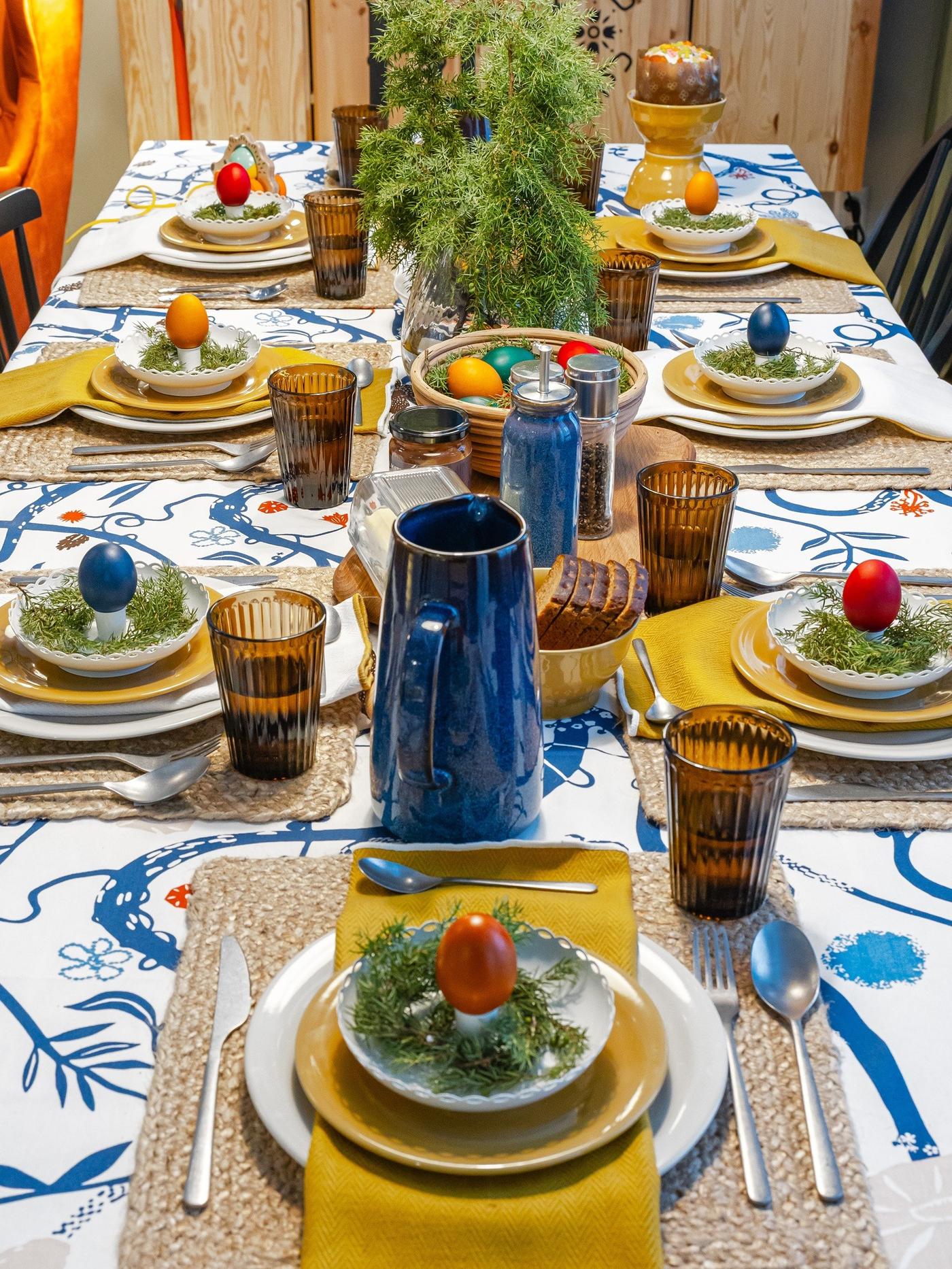 Table set with a vibrant precut fabric featuring blue floral motifs, styled with amber glasses, blue jugs and colourful eggs as playful decorations.