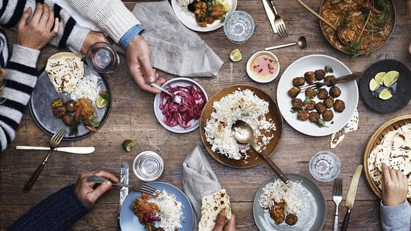 Table setting with meatballs, rice and pickles on a wooden table with cutlery and hands serving.