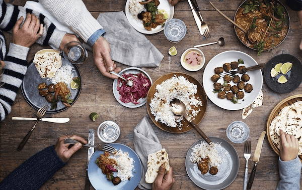 Table setting with meatballs, rice and pickles on a wooden table with cutlery and hands serving.
