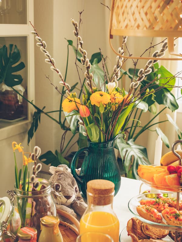 Table with a green vase of yellow and orange flowers, glassware, and plates of food in a bright, cozy dining space.