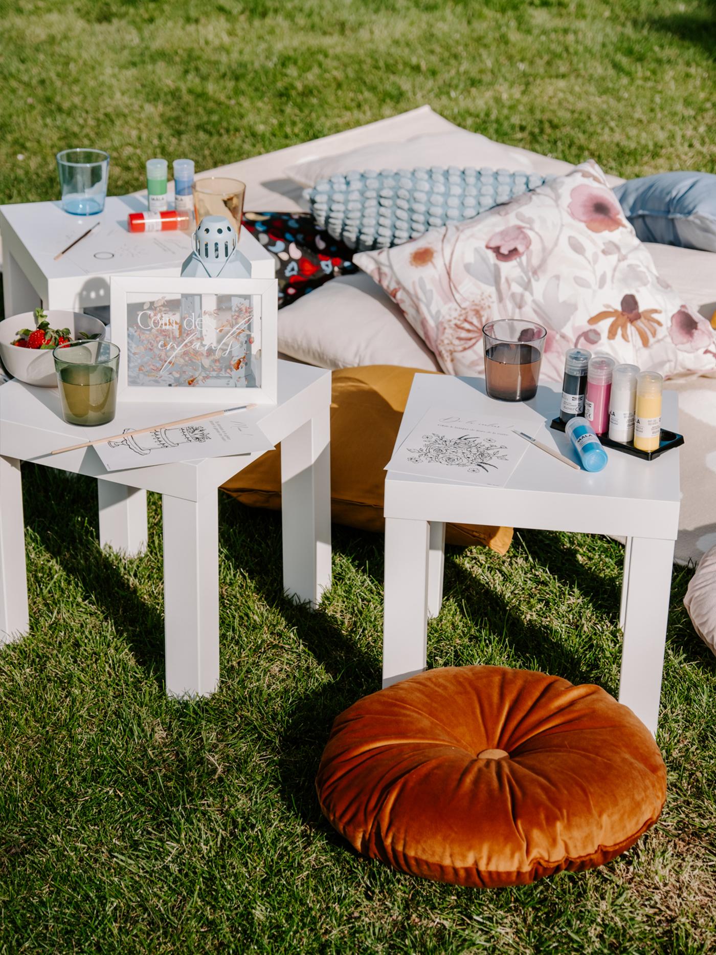 Tables blanches posées sur l'herbe avec des coussins colorées et des plaids pour une activité créative manuelle.