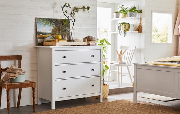 Tágas hálószoba, főleg fából készült bútorokkal és növényekkel, háromfiókos szekrénnyel.