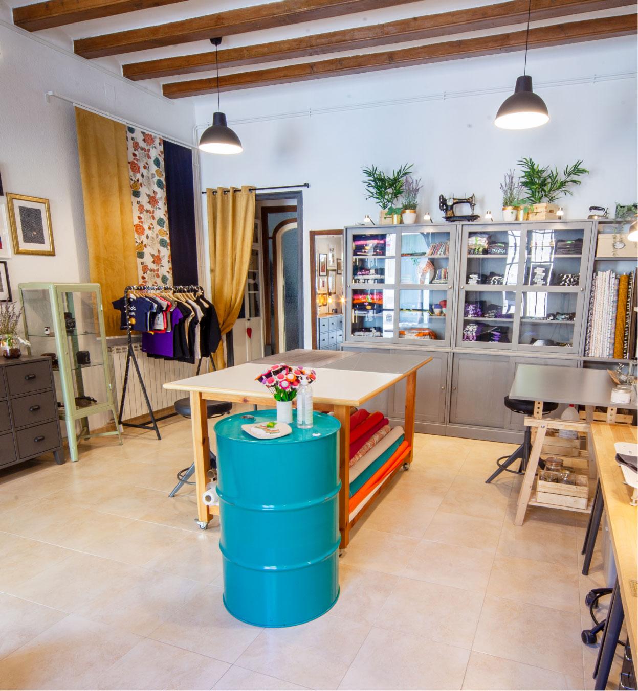 Taller creatiu amb taules de treball, roba i materials de costura, amb un ambient acollidor i decoració amb plantes i llums penjants.