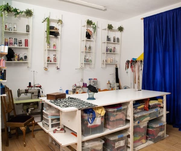 Taller de costura abans de ser reformat amb una gran taula de treball al centre, prestatgeries plenes d'objectes i eines de costura i una màquina de cosir antiga amb una butaca.