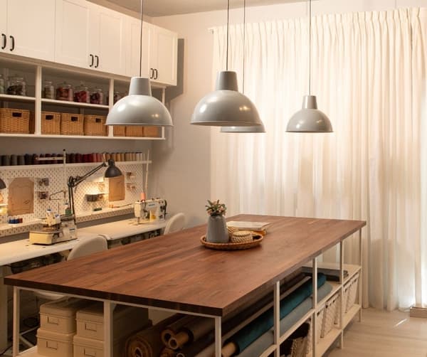 Taller de costura reformado con mesa ordenada, lámparas, estanterías y espacio de trabajo.