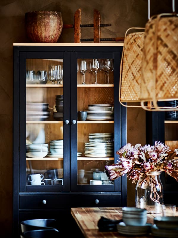 Tamna HEMNES vitrina ispunjena posuđem stoji uz zid koji se nalazi pokraj blagovaonskog stola.