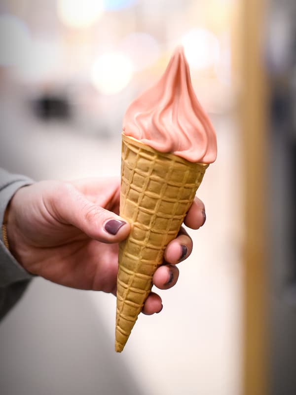 Tangan seseorang memakai baju berwarna kelabu dan penggilap kuku warna ungu sedang memegang kon aiskrim diisi dengan ais lembut strawberi.