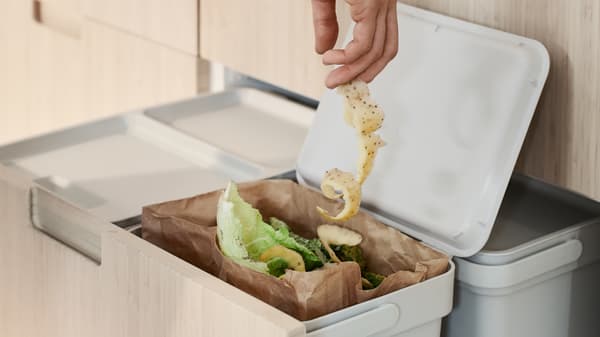 Tangan seseorang sedang memasukkan seuntai panjang kulit ubi kentang ke dalam tong HÅLLBAR yang mengandungi sisa dapur organik.