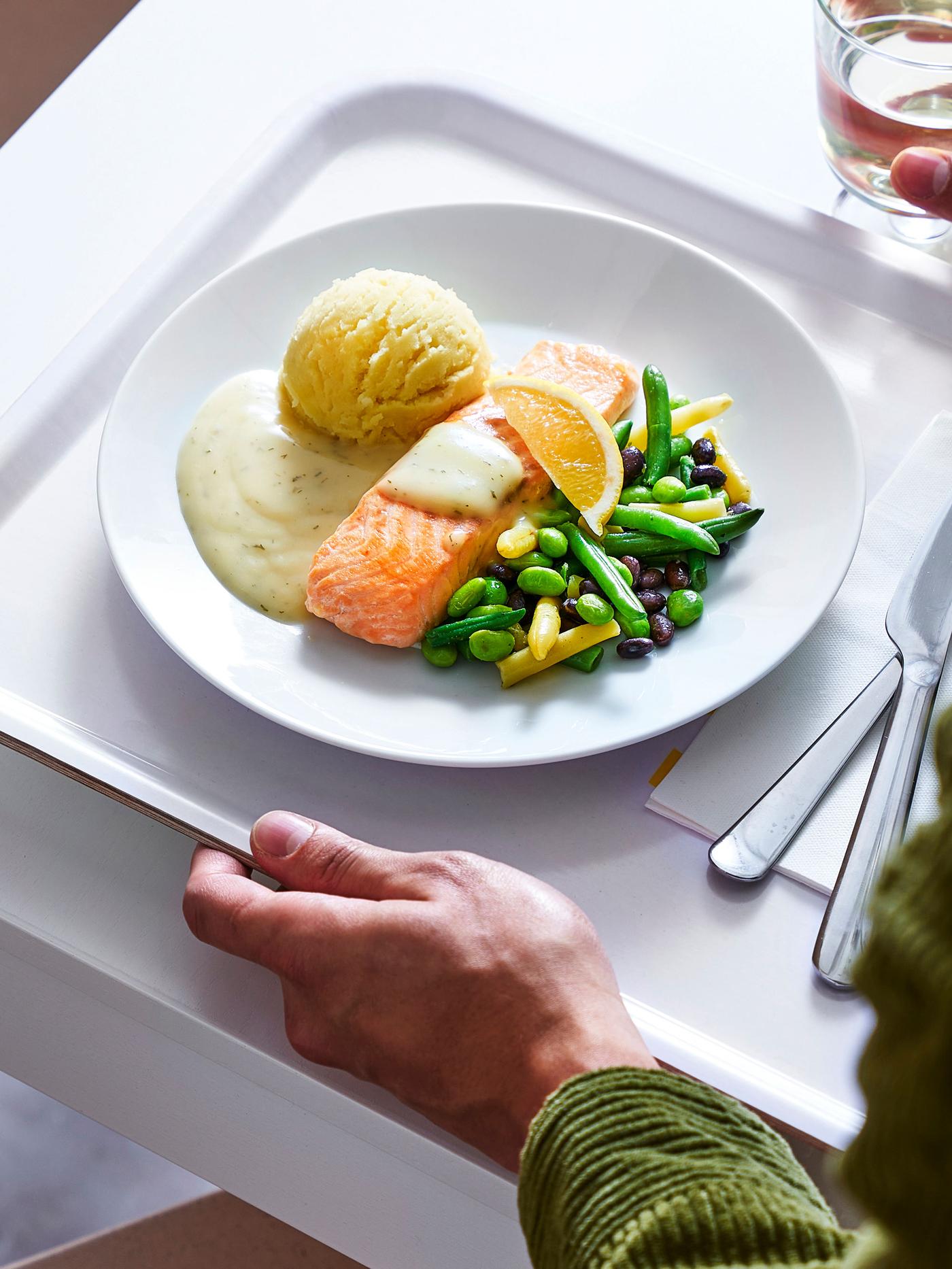 Tanji pladanj na bijelom stolu s tanjurom hrane: pečeni losos, pire krumpir, povrće (zelene mahune, grašak, mrkva) i pecivo. Uz pladanj su pribor za jelo i čaša vode.