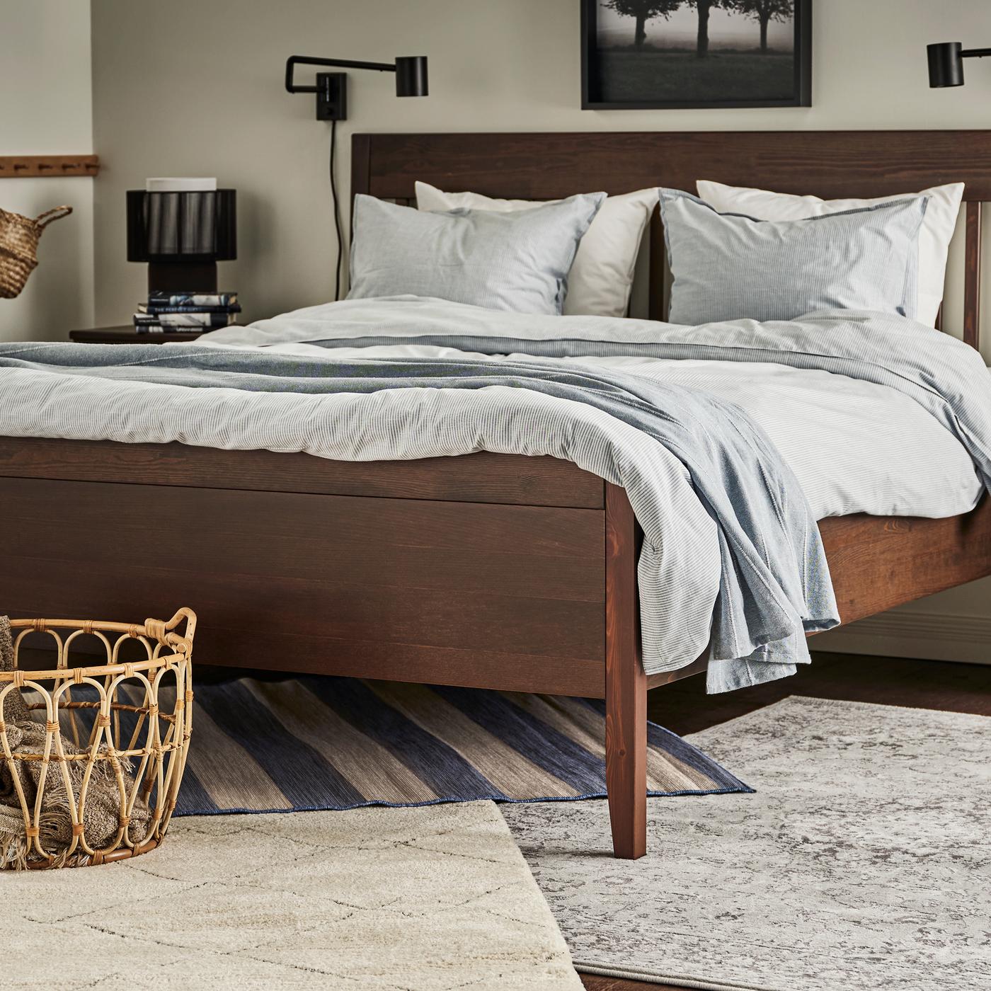 Tapetes VRENSTED, VEDBÄK e TVERSTED debaixo de uma cama IDANÄS em castanho escuro, feita com roupa de cama BERGPALM em azul/às riscas e manta em cinzento.