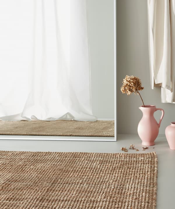 Tapis en jute sur un sol gris, un vase rose avec des fleurs séchées posé à côté d’un grand miroir blanc dévoilant un rideau blanc.