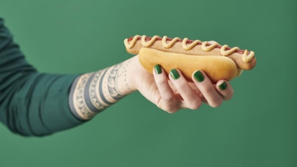 Tattooed arm holding a hot dog with mustard against a solid green background.