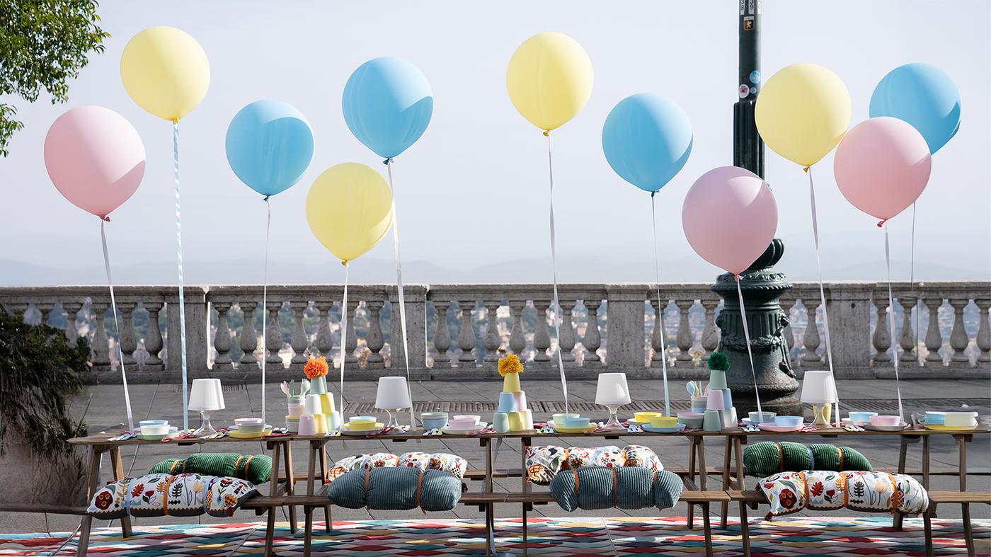 Tavola all’aperto colorata con cuscini e palloncini, pronta per una festa speciale all’insegna della convivialità e dello spirito IKEA.