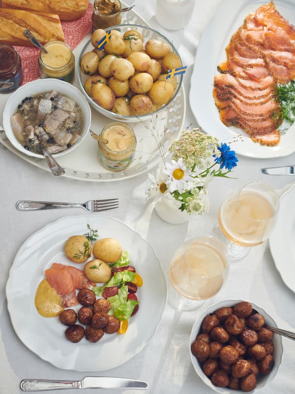 Tavola con piatti, posate ÄTBART, salmone marinato su un piatto, polpette in una ciotola e aringhe su un vassoio ROMANTISK - IKEA
