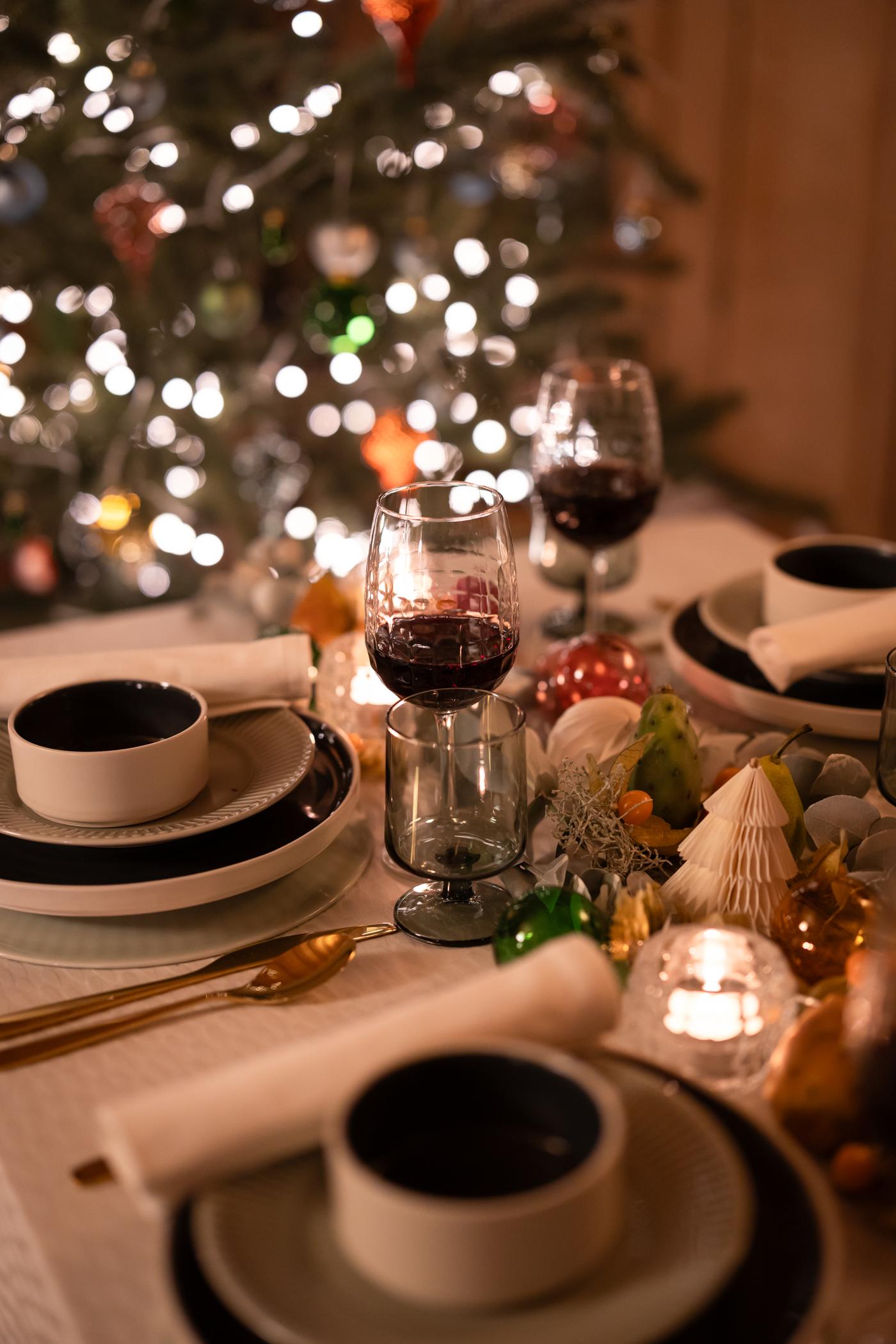 Tavola imbandita a festa con eleganti stoviglie, bicchieri da vino, posate dorate e decorazioni natalizie; un albero di Natale illuminato sullo sfondo aggiunge calore.