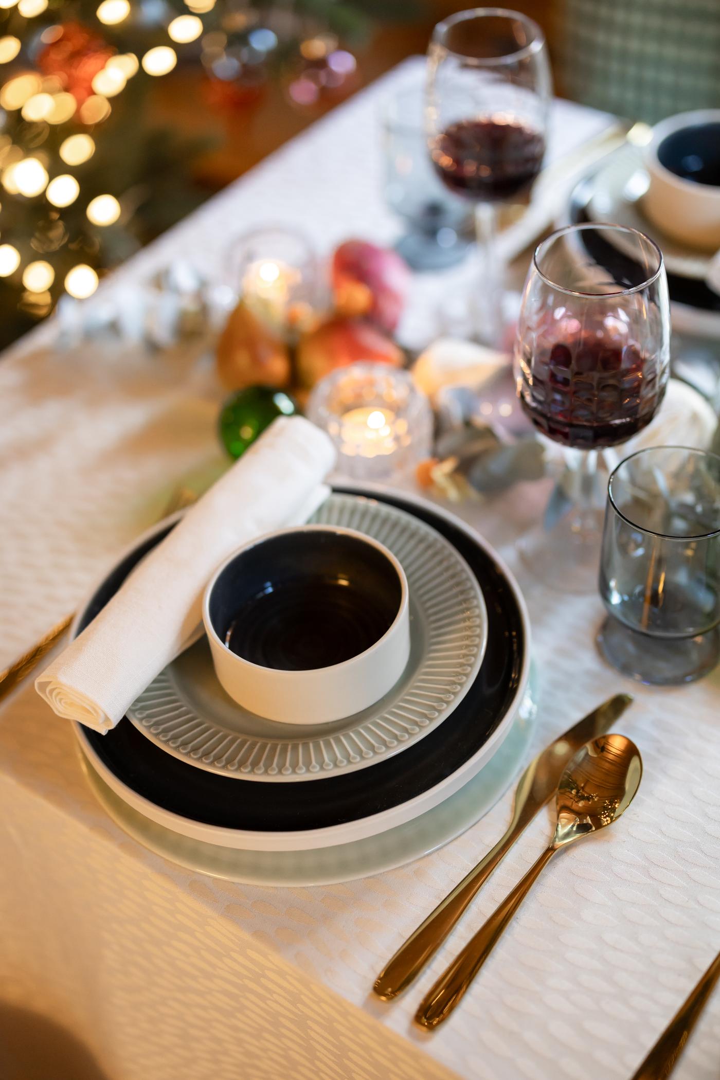 Tavola imbandita per le feste con elegante apparecchiatura: piatti impilati, una ciotola, tovaglioli piegati, posate dorate, bicchieri da vino e morbida luce di candela. Accogliente atmosfera festiva.
