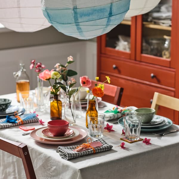 Tavolo con tovaglia color naturale/beige, apparecchiato con piatti spaiati di vari colori, bicchieri POKAL e fiori artificiali.