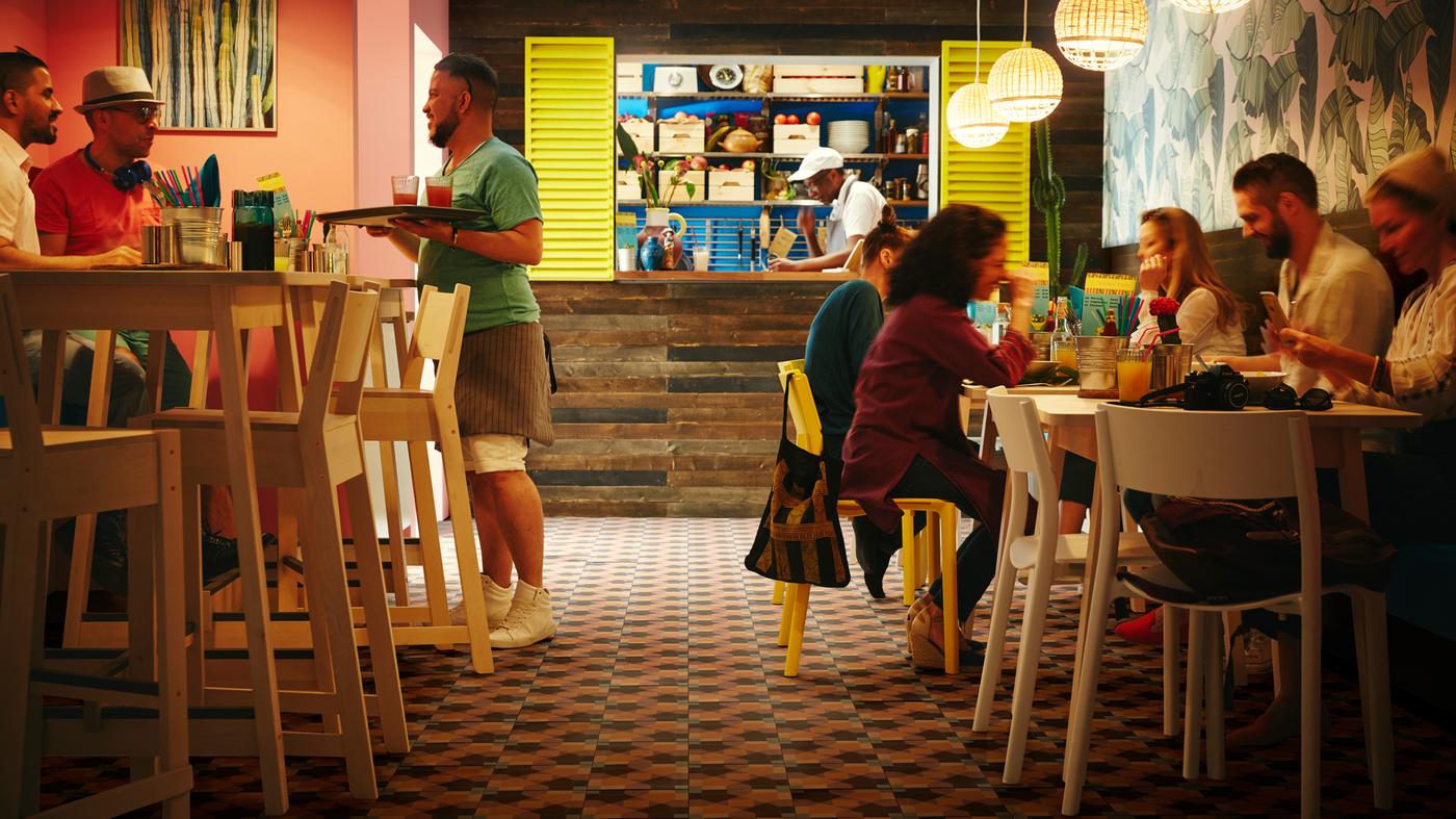 طاولات مرتفعة NORRÅKER بيضاء من خشب البتولا ومقاعد مرتفعة من خشب البتولا في مطعم صغير مع مصابيح معلقة من الخيزران وبعض العملاء.