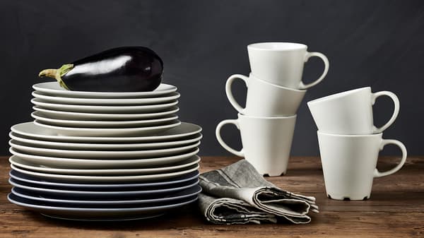 Tazze DINERA beige impilate su un piano di lavoro, vicino a una pila di piatti beige e grigio-blu di diverse dimensioni - IKEA
