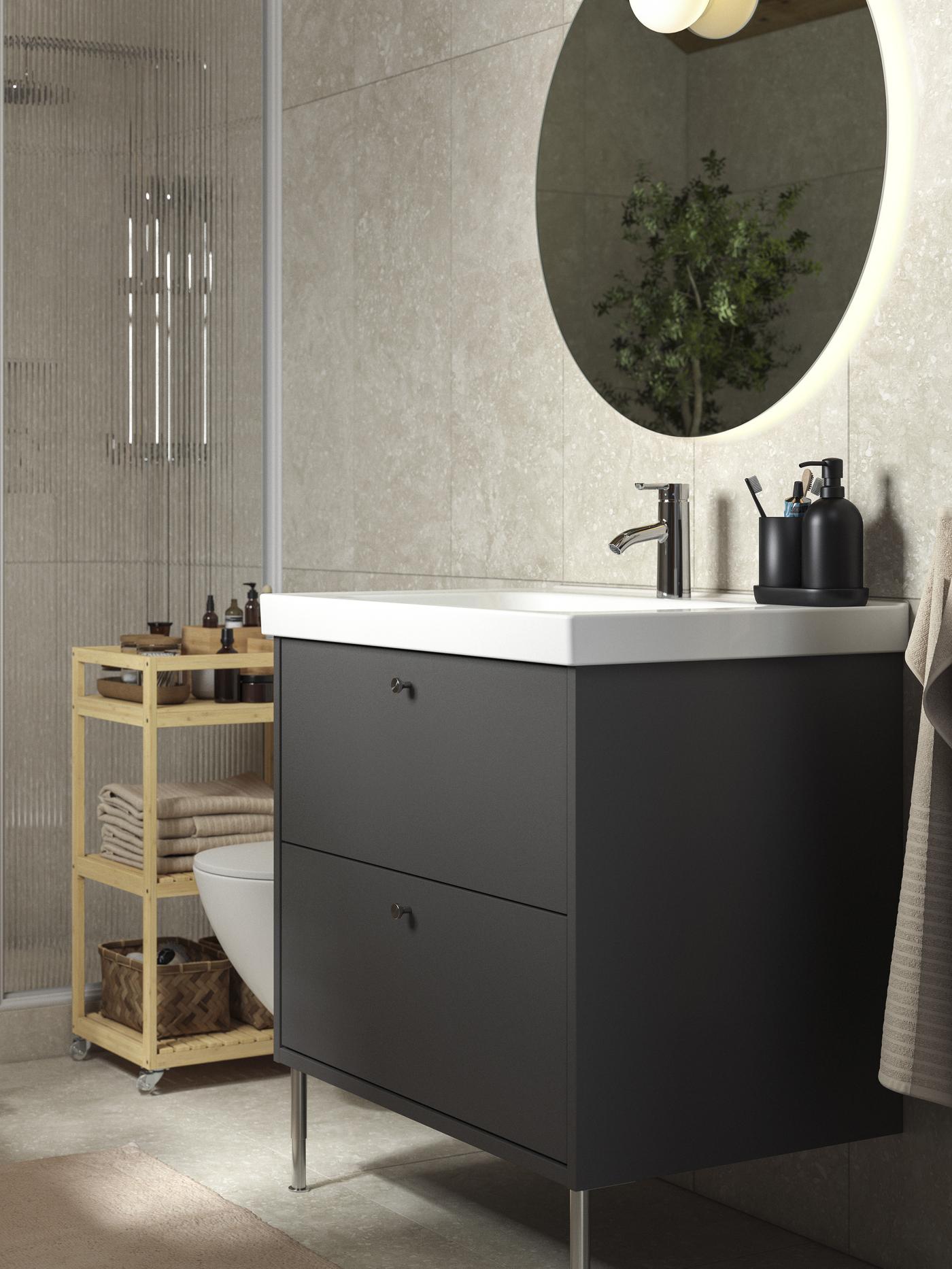 Tchnąca spokojem, oświetlona naturalnym światłem łazienka z ciemnoszarą szafką HAVBÄCK z szufladami i meblami z naturalnego bambusa. W kącie stoi duża zielona roślina.