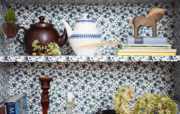 Teapots, books and ornaments on shelves, which are covered with a blue floral fabric.