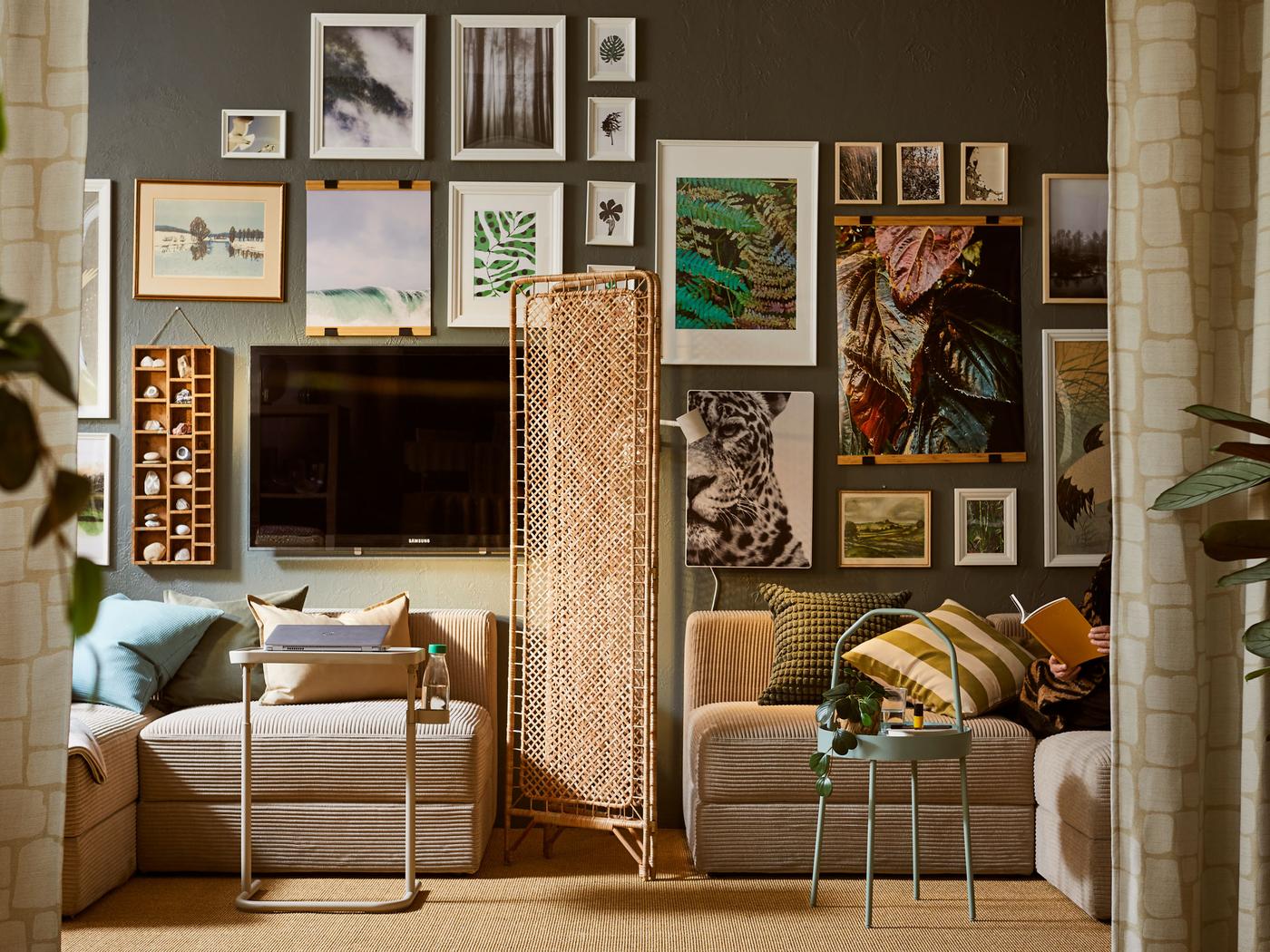 Teile eines JÄTTEBO Sofas stehen auf beiden Seiten eines TOLKNING Raumteilers aus Rattan. Dahinter ist eine Wand mit gerahmten Bildern zu sehen.