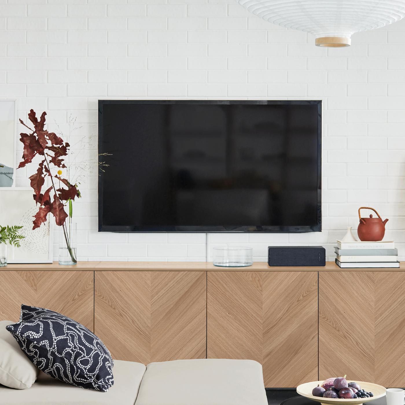 Téléviseur noir fixé sur un mur blanc au-dessus d’une combinaison de rangement BESTÅ avec des façades de portes HEDEVIKEN en plaqué chêne.