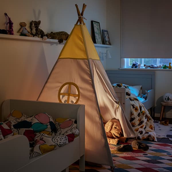 Tenda de brincar HÖVLIG sobre tapete BUSENKEL multicor, entre duas camas extensíveis em branco. Prateleiras para molduras na parede.