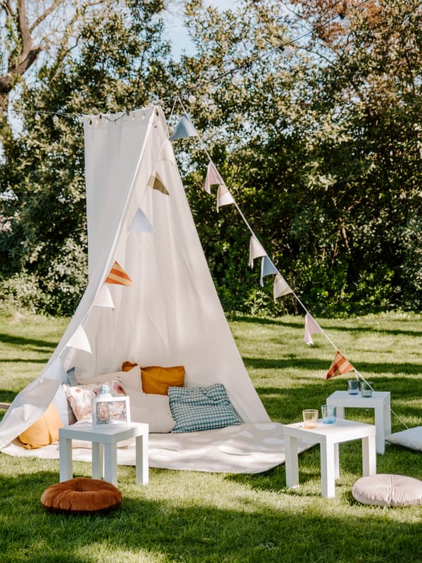 Tente faite à partir de draps blancs accrochés à un arbre dehors avec des coussins et un plaid et quelques tables blanches.