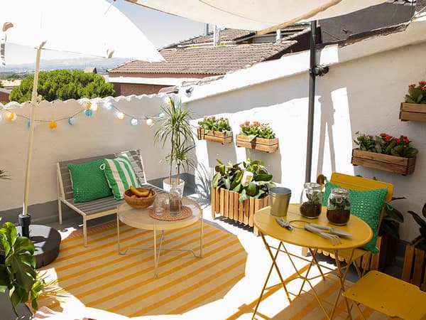 Terrassa amb tauletes, cadires, catifes, un sofà, una garlanda de llums, portatestos de fusta amb plantes, un para-sol i estris de jardineria.